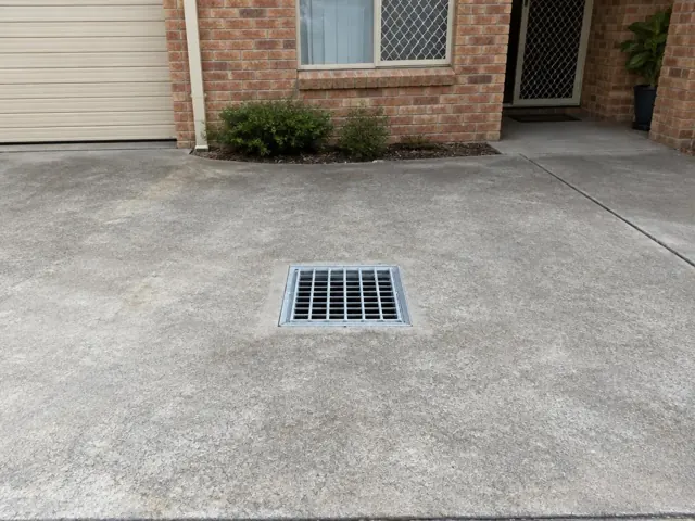 Clean metal stormwater grate set in a concrete driveway beside a brick house in Kings Langley