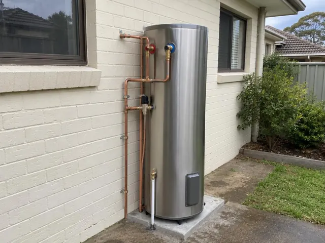 Stainless steel hot water cylinder with copper pipework beside a painted brick house wall in Parklea